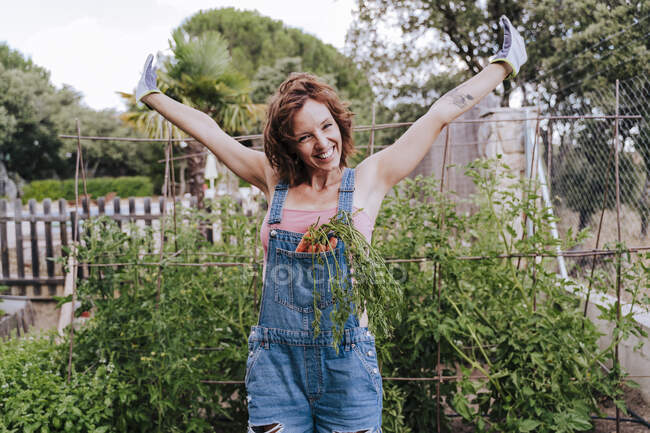 Mujer alegre con los brazos levantados de pie contra las plantas en huerta - foto de stock