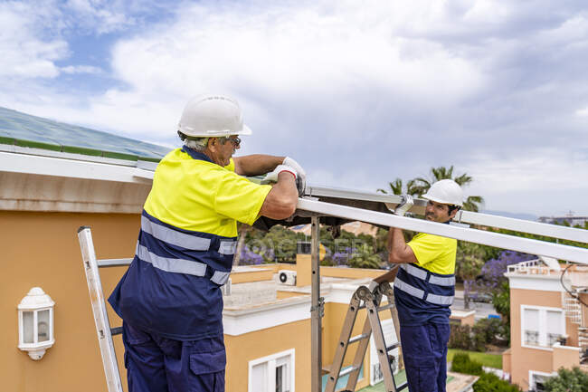 Compañeros de trabajo masculinos instalando paneles solares en el techo de la casa mientras están de pie sobre escaleras contra el cielo nublado - foto de stock