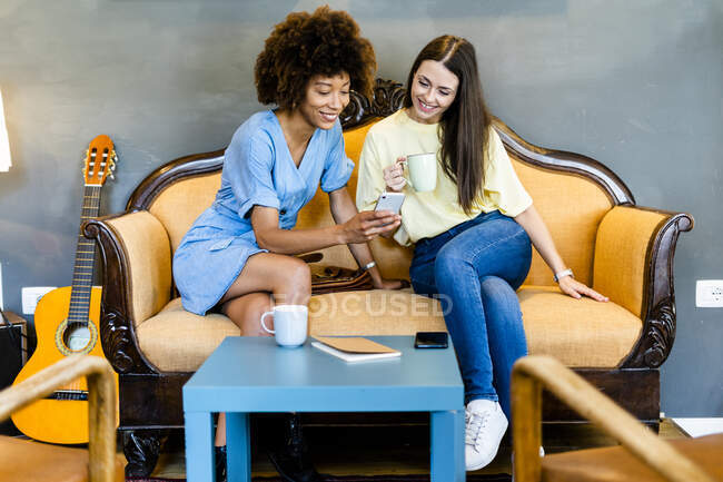 Мультиетнічні жінки-друзі користуються смартфоном, сидячи на дивані в сучасній кафе. — стокове фото