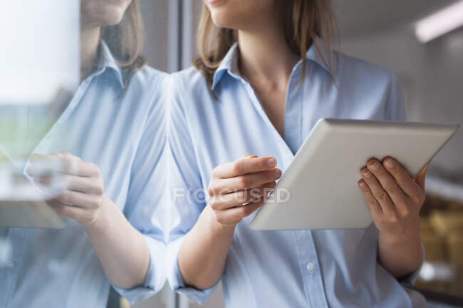 Mujer adulta que usa tableta digital mientras está de pie junto a la ventana en casa - foto de stock