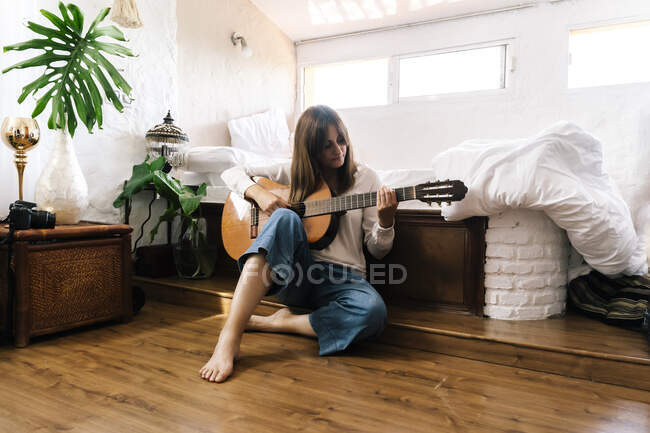 Woman sitting on the floor in front of bed playing guitar — Stock Photo