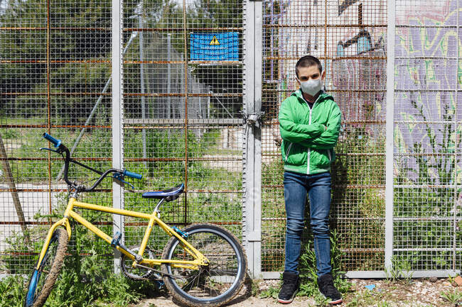 Ritratto di ragazzo pre-adolescente con maschera facciale in piedi con le braccia incrociate in bicicletta contro la recinzione — Foto stock