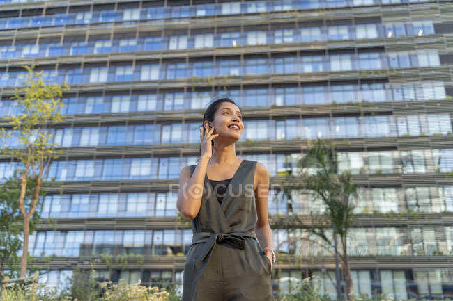 Sorridente donna d'affari che parla tramite auricolare wireless mentre in piedi contro edificio in città — Foto stock