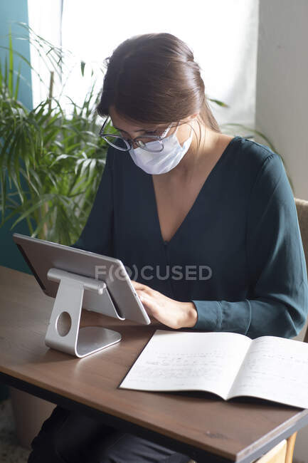 Mujer joven con máscara protectora usando tableta en el escritorio en casa - foto de stock