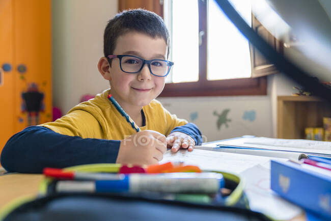 Улыбающийся студент, сидящий за столом с книгами во время коронавируса — стоковое фото