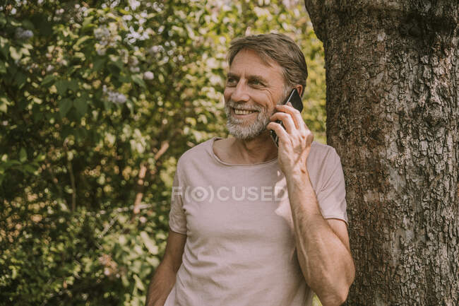 Lächelnder älterer Mann telefoniert im Garten an Baum gelehnt — Stockfoto
