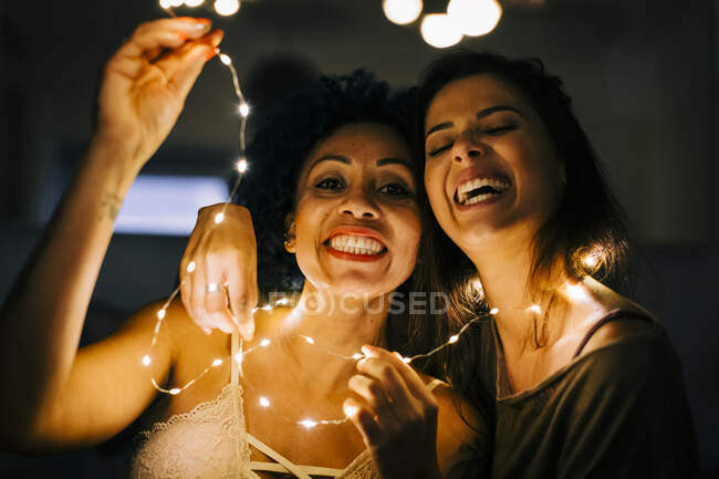 Primo piano di amiche allegre che tengono luci di stringa illuminate a casa — Foto stock