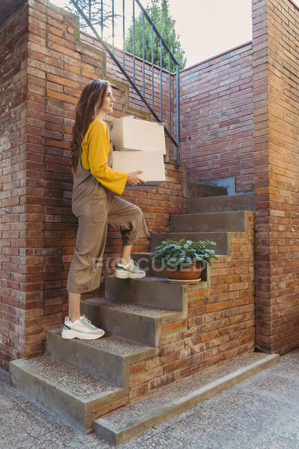 Jeune femme portant des boîtes blanches tout en se déplaçant sur les marches par un mur de briques — Photo de stock