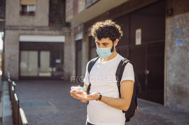 Joven con máscara lavándose las manos con desinfectante mientras está parado contra la construcción - foto de stock