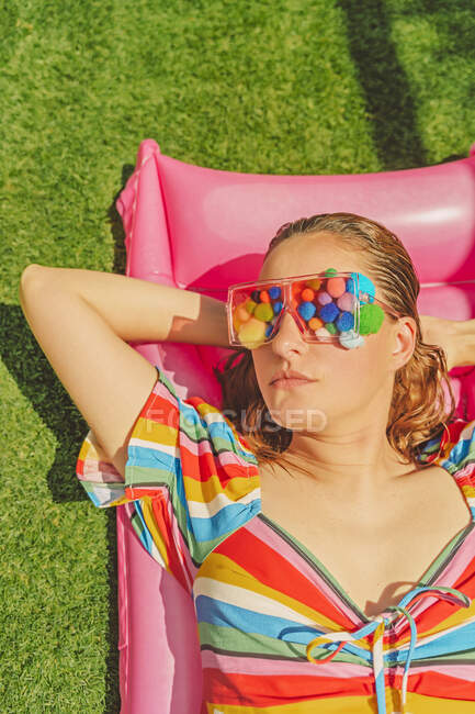 Retrato de mulher usando óculos com pompons coloridos cobrindo os olhos relaxando no leito de ar rosa — Fotografia de Stock
