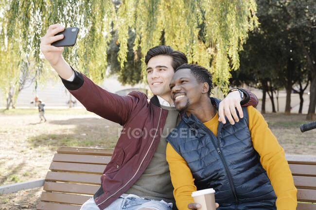 Deux jeunes hommes heureux assis sur le banc du parc prenant un selfie — Photo de stock
