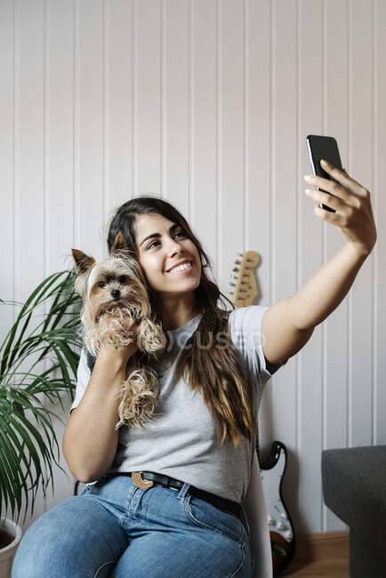 Jovem mulher tomando selfie com Yorkshire Terrier enquanto se senta na cadeira em casa — Fotografia de Stock