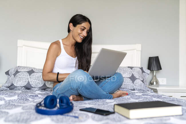Усмішка за допомогою ноутбука сидячи з книжкою та навушниками на ліжку. — стокове фото