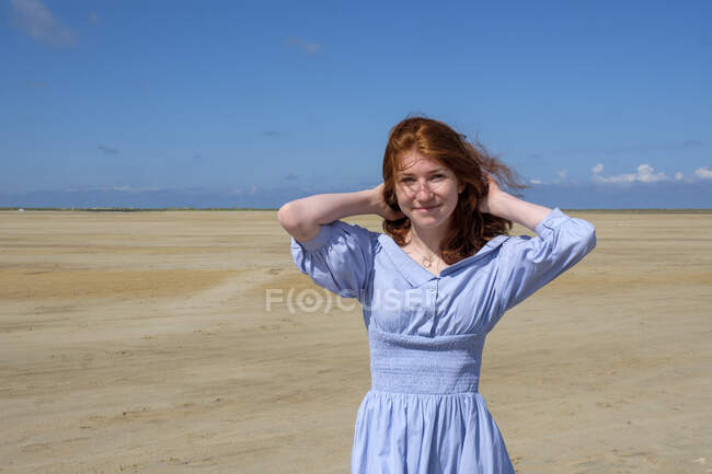 Retrato de una adolescente sonriente con vestido azul de pie en la playa contra el cielo en un día soleado - foto de stock