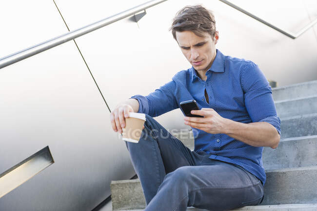 Uomo d'affari in possesso di caffè utilizzando smartphone mentre seduto su gradini — Foto stock