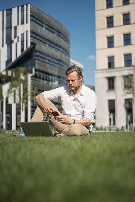 Uomo d'affari con computer portatile seduto in erba in città utilizzando smartphone — Foto stock