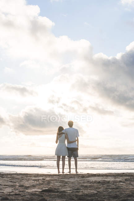 Amour jeune couple avec les bras autour de regarder la mer contre ciel nuageux — Photo de stock