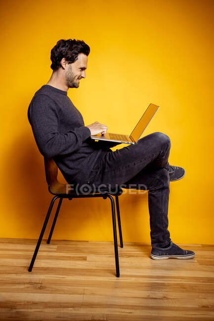 Hombre sonriente usando el ordenador portátil mientras está sentado en la silla por el fondo amarillo - foto de stock