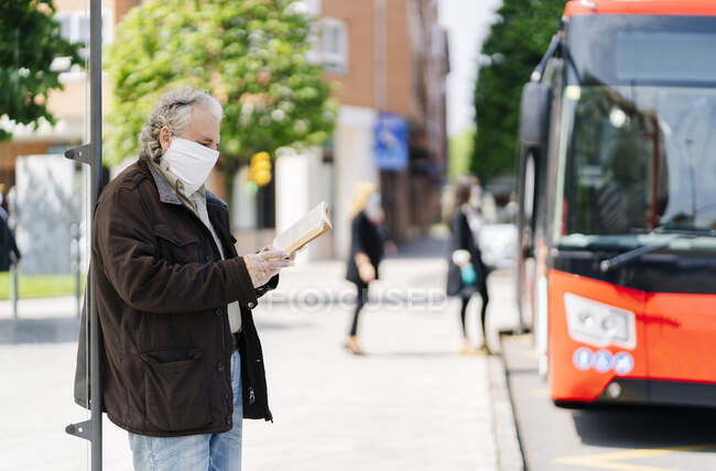 Старший мужчина в защитной маске и перчатках читает книгу во время ожидания на автобусной остановке, Испания — стоковое фото