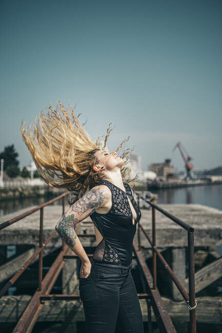 Joven mujer tatuada lanzando su largo cabello rubio mientras está de pie en el muelle contra el cielo despejado durante el día soleado - foto de stock