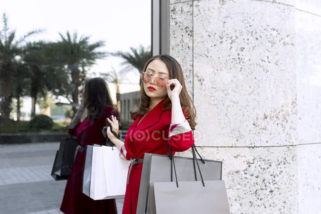 Молодая женщина в солнечных очках носит сумки с покупками, стоя рядом с современным зданием — стоковое фото