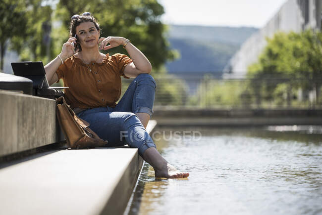 Mujer sonriente hablando por teléfono móvil mientras está sentada junto al estanque en el parque durante el día soleado - foto de stock