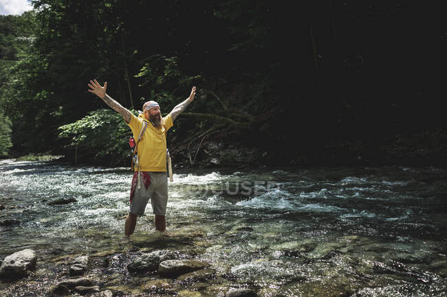 Уокер с бородой и желтой толстовкой, стоящий в горной реке и поднимающий руки — стоковое фото