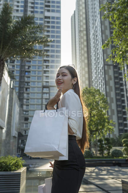 Smiling young woman holding shopping bags while standing against modern buildings in city — Stock Photo