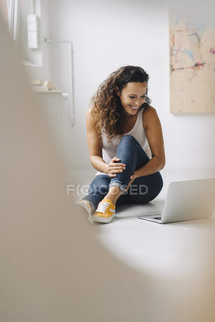Усміхнена жінка дивиться відео через ноутбук, сидячи вдома на підлозі. — стокове фото