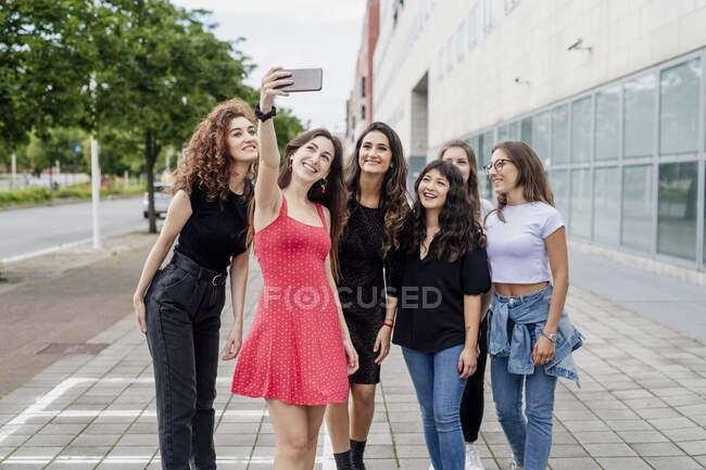 Smiling young woman taking selfie with female friends while standing on footpath in city — Stock Photo