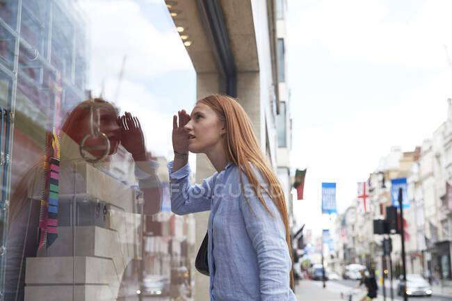 Redhead woman shielding eyes while looking through store window in city - foto de stock