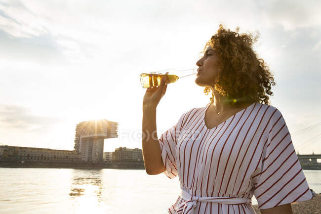 Mid adult woman with curly hair drinking beer while standing against sky in city at sunset — Stock Photo