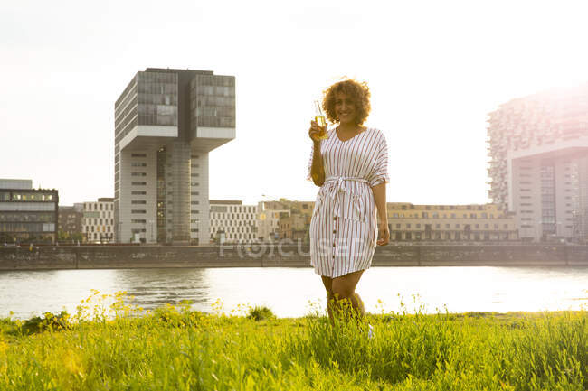 Smiling woman holding beer bottle while standing on grassy land against river in city at sunset — Fotografia de Stock