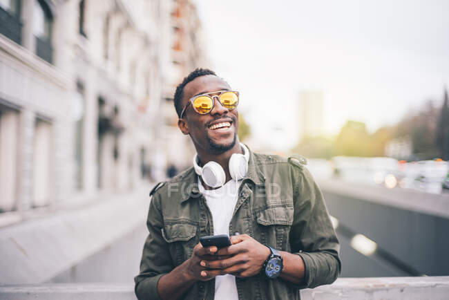 Hombre alegre con gafas de sol usando el teléfono inteligente mientras está de pie en la ciudad - foto de stock