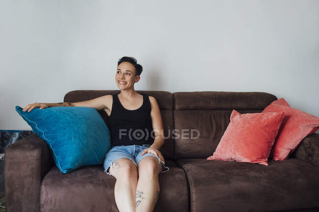 Mujer sonriente mirando hacia otro lado mientras se sienta en el sofá - foto de stock