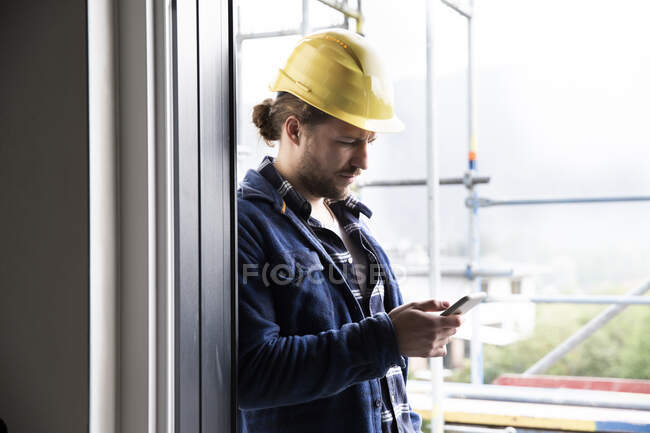 Construction worker wearing helmet using smart phone by window at constructing house — Stock Photo