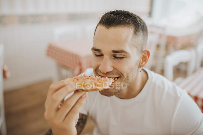 Primer plano del joven con los ojos cerrados comiendo pizza mientras está sentado en el restaurante - foto de stock