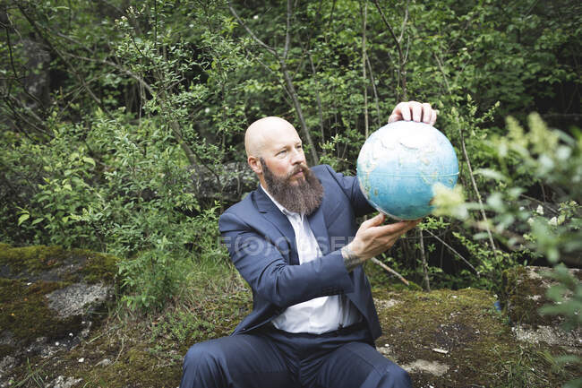 Homem de negócios barbudo careca segurando globo enquanto sentado contra árvores na floresta — Fotografia de Stock