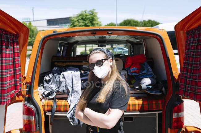 Donna con maschera viso e occhiali da sole in piedi davanti al furgone aperto con le braccia incrociate — Foto stock