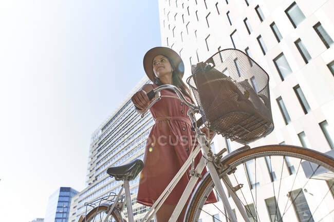 Задумчивая женщина, стоящая с велосипедом на фоне зданий и неба — стоковое фото