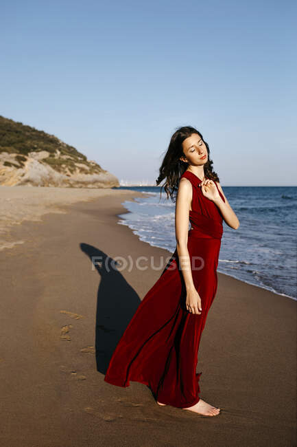 Zarte Frau in rotem Kleid, die am Meer steht und die Sonne spürt — Stockfoto