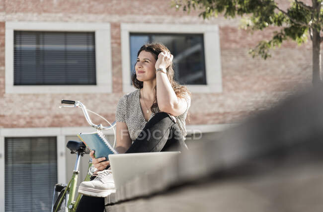 Улыбающаяся женщина, держащая в руках книгу, размышляет, сидя против строительства в городе — стоковое фото