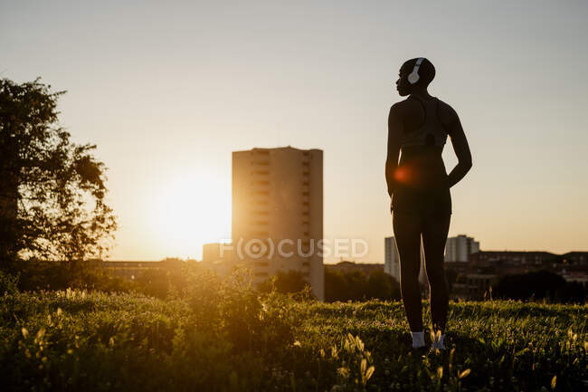 Silhouette femme écouter de la musique tout en se tenant sur la terre herbeuse contre le ciel clair pendant le coucher du soleil — Photo de stock