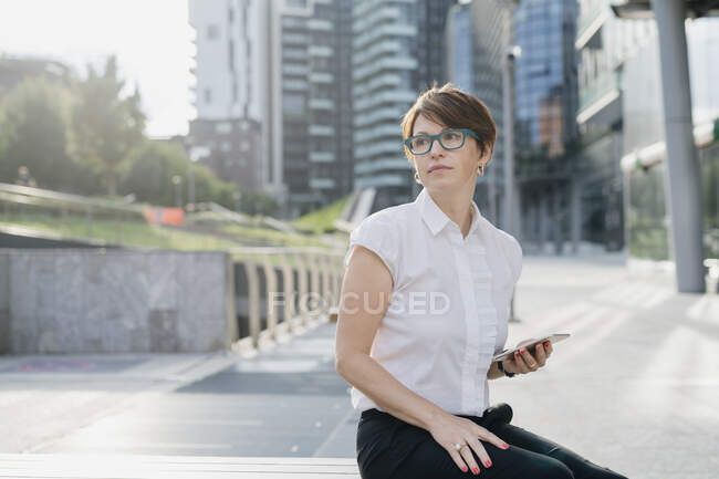 Thoughtful businesswoman holding smart phone while sitting against building in city - foto de stock