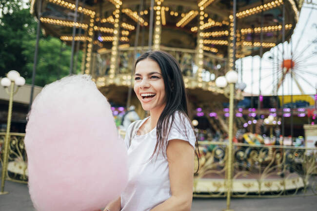 Cheerful young woman with cotton candy standing in amusement park — Fotografia de Stock