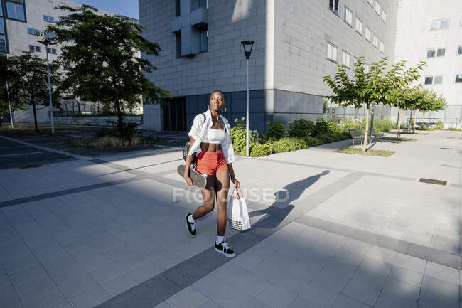 Молодая женщина с сумкой и скейтборд ходить по тропинке в городе — стоковое фото