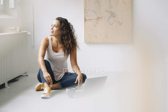 Mujer reflexiva con portátil en el suelo mirando hacia otro lado mientras está sentado en casa - foto de stock