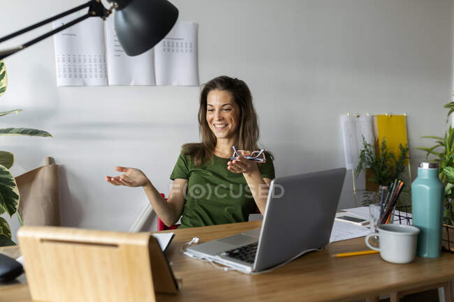 Mujer joven sonriente haciendo gestos mientras mira la tableta digital en el escritorio en la oficina en casa - foto de stock