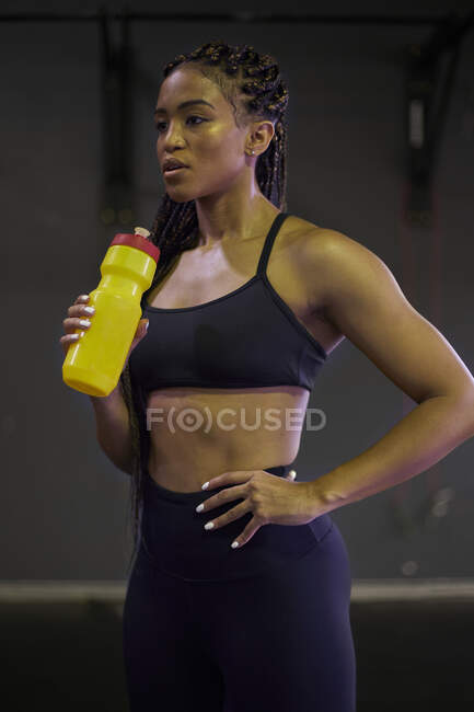Atleta femenina reflexiva sosteniendo la botella de agua mientras está de pie en el gimnasio - foto de stock