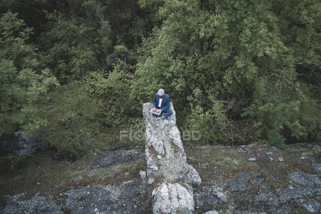 Vista aérea del hombre de negocios que usa traje usando el ordenador portátil mientras está sentado en la roca en el bosque - foto de stock